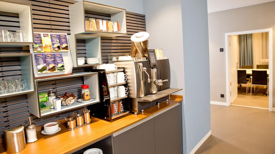 Office kitchenette with a coffee machine, assorted coffee supplies, mugs, and shelves with tea boxes. A meeting room is visible in the background.