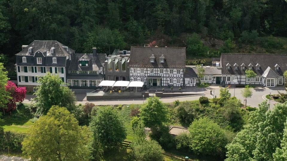 Aerial view of a large, historic building complex surrounded by trees, featuring white walls and dark patterned woodwork.