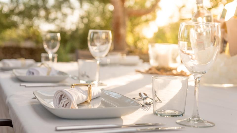 Elegant outdoor dining table set with white plates, glasses, napkins, and cutlery in soft, natural lighting.