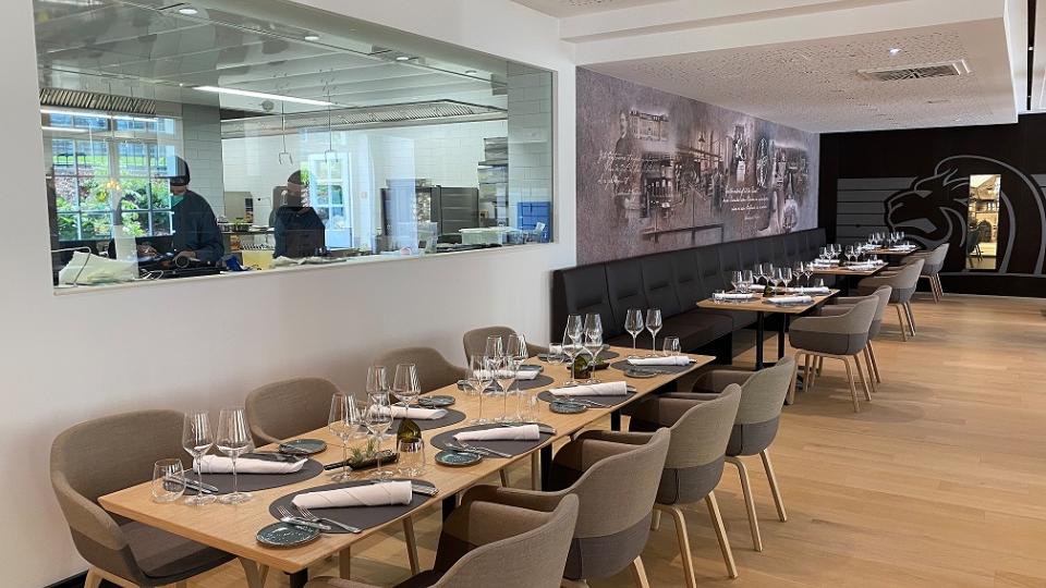 A modern restaurant interior with neatly set tables, chairs, and wine glasses. A glass panel reveals the kitchen where chefs are at work. The decor is minimalist with a wooden floor.